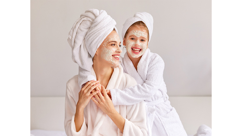 Two people in robes and towels with facial masks, smiling at each other.